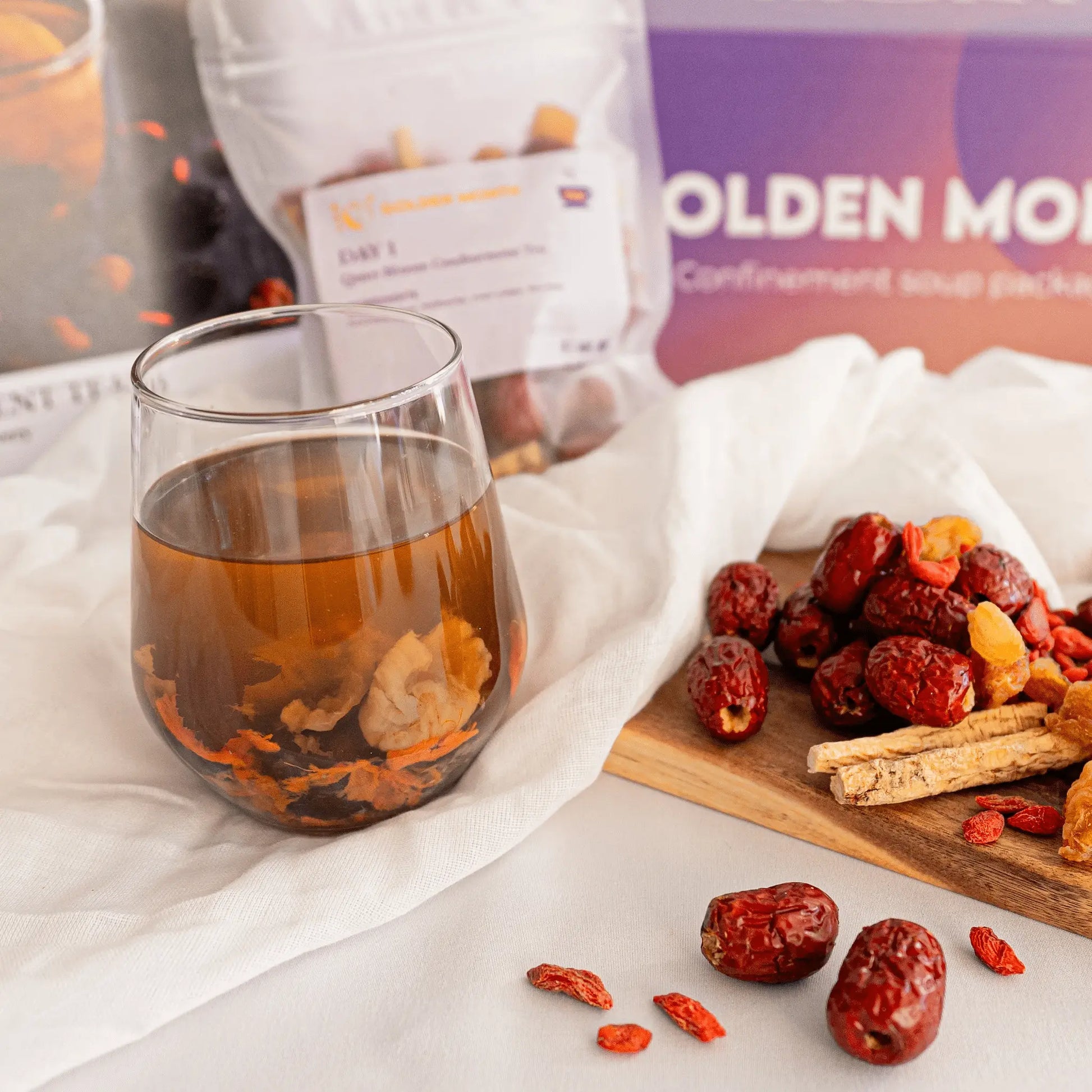 Glass of postpartum herbal tea with dried dates and a package labeled 'Golden Month' on a white surface.