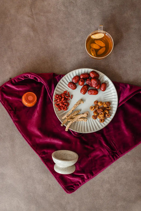 Tea cup with dried lactation herbs and roots on a plate, placed on a maroon fabric with a brown background.