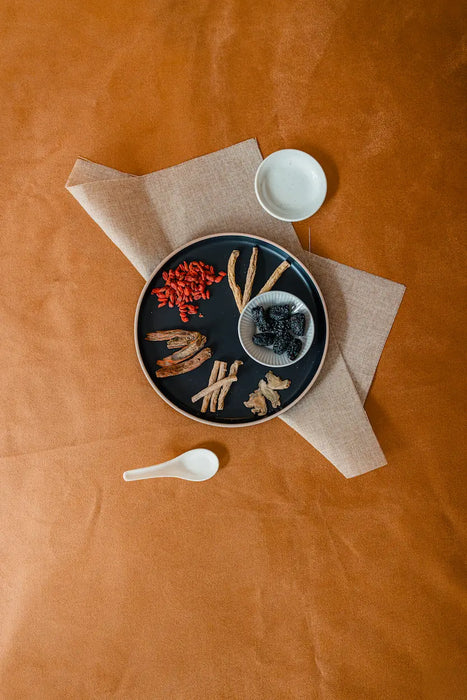 Dried confinement soup herbs on a black plate with a white bowl and spoon on a brown surface
